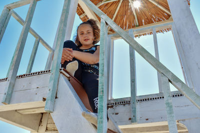 Low angle view portrait of girl sitting on staircase of lifeguard hut