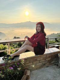 Portrait of woman sitting on retaining wall against sky during sunset