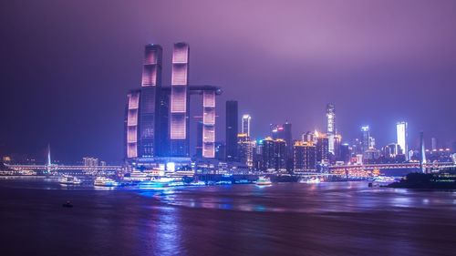 Illuminated modern buildings by bay against sky at night