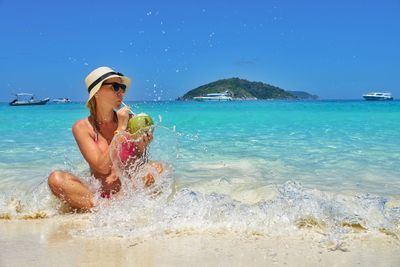 Woman drinking coconut water while sitting at beach against blue sky