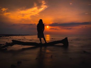 Full length of man standing on driftwood at beach against sky during sunset