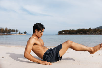 Rear view of shirtless man sitting at beach against sky