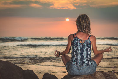 Woman doing yoga on rock at beach against sky during sunset