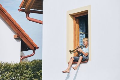 Boy playing trumpet while sitting on window of house