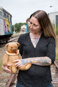 A young caucasian girl with glasses and tattoos smiles, a brown teddy bear