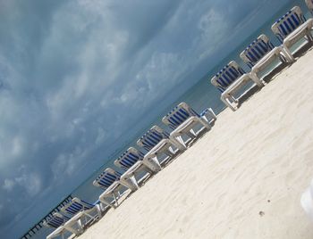 Tilt image of deck chairs at beach against cloudy sky