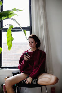 Young woman looking away while sitting on window at home