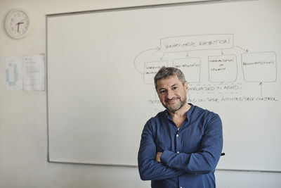 Portrait of confident male teacher standing with arms crossed in front of whiteboard at university classroom