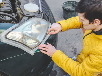A guy in a yellow jacket sticks scoich around the edges of the headlight preparing it for polishing