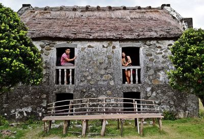 Low angle view of man and woman by window of old house by trees