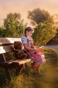 Full length of girl reading book while sitting on bench during sunset