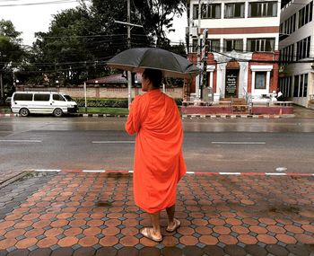 Rear view of monk with umbrella standing on sidewalk in city