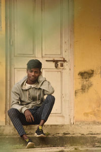 Full length portrait of young man sitting at entrance of building