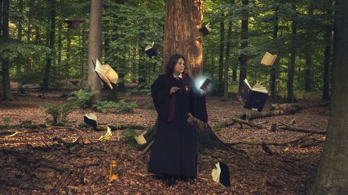 Teenage girl holding magic wand while standing amidst books at forest