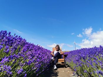 Full length of woman sitting on chair amidst flowering plants against blue sky