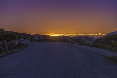 Empty road by illuminated city against clear sky at night