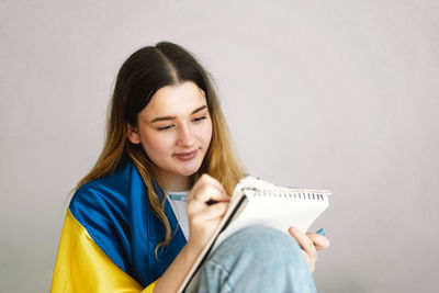 A ukrainian teenage girl is wrapped in the ukrainian flag and is drawing or doing homework