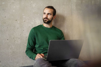 Thoughtful businessman with laptop sitting in front of wall at office