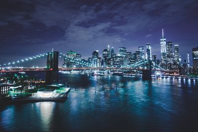 Illuminated bridge over river by buildings against sky at night