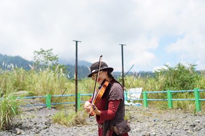 Woman playing violin while standing on field against cloudy sky