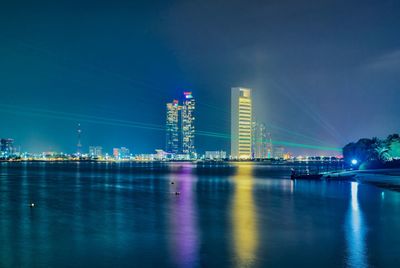 Illuminated buildings by river against blue sky at night