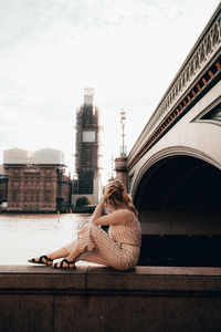 Woman sitting on retaining wall by bridge in city against sky