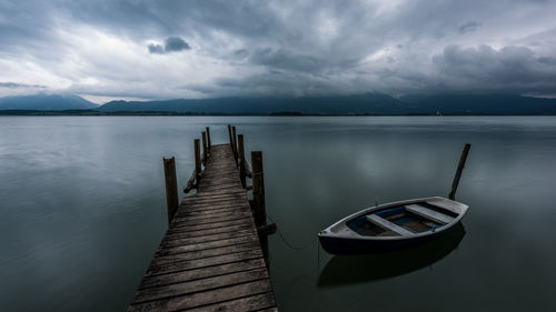 Boat moored by wooden pier in lake against cloudy sky