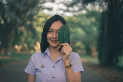 Portrait of smiling young woman holding leaf while standing at public park