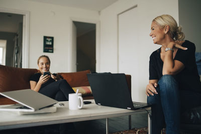 Smiling teenage girl and mother sitting with laptops on coffee table in living room at home