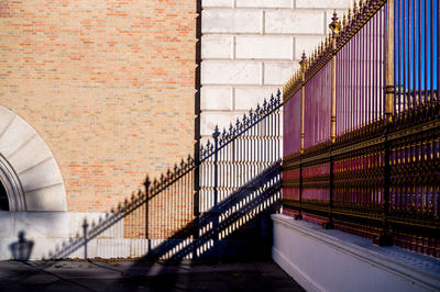 A tall old fence with uprights and spikes casts a clear shadow on the façade of a historic building.