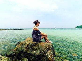 Side view of young woman sitting on rock by sea against sky