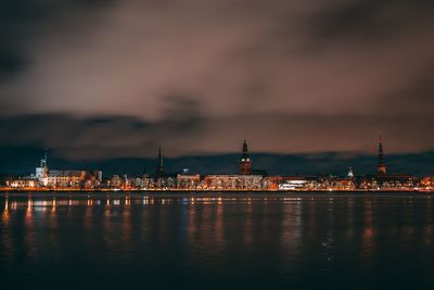 Illuminated city at waterfront against cloudy sky