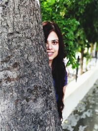 Portrait of smiling young woman looking from behind tree trunk