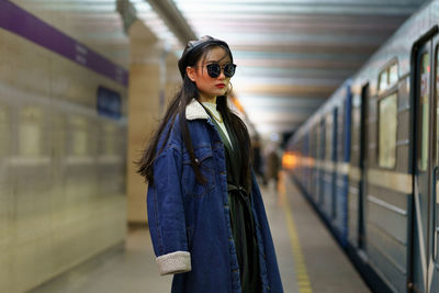 Stylish asian girl in glasses and street fashion outfit at underground platform with arriving train