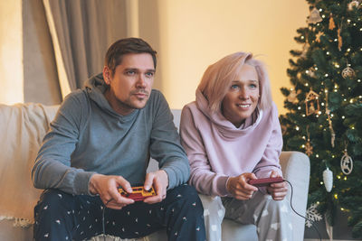 Fun young family in home clothes playing video games on couch on background of christmas tree