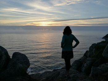 Rear view of woman with hands on hip standing on rocky beach during sunset