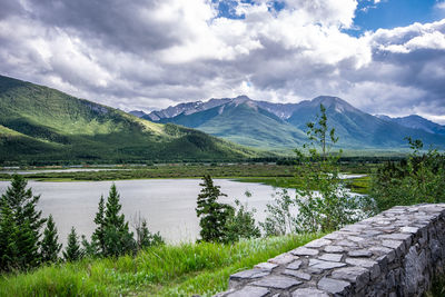 Summer scenic view with stone wall, grass, trees, lake, train, mountains, cloudy blue sky background