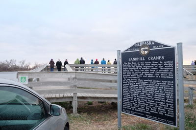 Car parked by historical marker with people on pier against sky