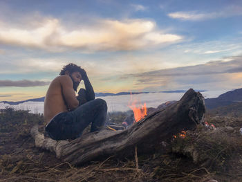 Side view of man sitting on shore against sky during sunset