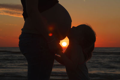 Silhouette son kissing pregnant mother at beach against sky during sunset