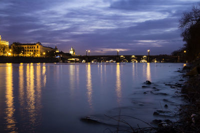 Buildings lit up at night at lakeshore against cloudy sky