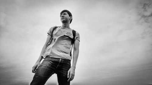 Low angle view of young man looking away while standing against cloudy sky
