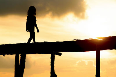 Low angle view of silhouette girl walking on pier against orange sky