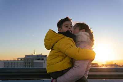 Woman with child in arms standing on rooftop with sunset light in background