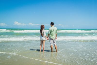 Rear view of couple holding hands while standing at seashore against sky