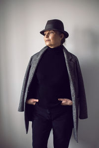 Fashionable older woman in black clothes and a hat stands against a white wall in the office