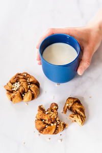 Female hand holding a blue cup of milk next to cinnamon rolls lying on a table at home.