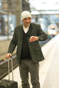 Smiling mature man with suitcase checking time at railroad station platform