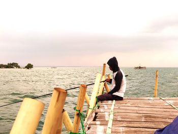 Side view of man fishing while sitting on pier in sea against sky