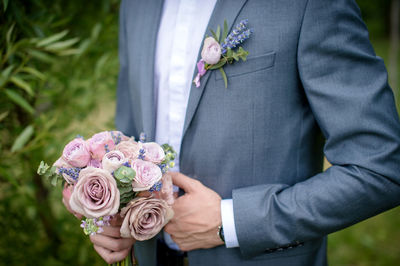 The groom stands with a wedding bouquet of roses and holds a jacket